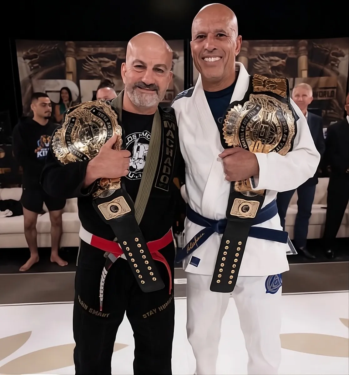 Carlos Machado and Royce Gracie standing together, each holding a Legends of Jiu-Jitsu belt
